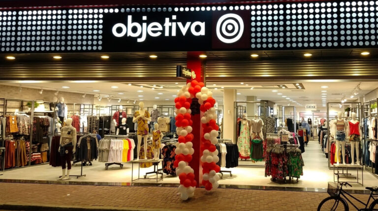Fachada iluminada da loja Moda Objetiva com letreiro em destaque, arco de balões na entrada e manequins no interior.